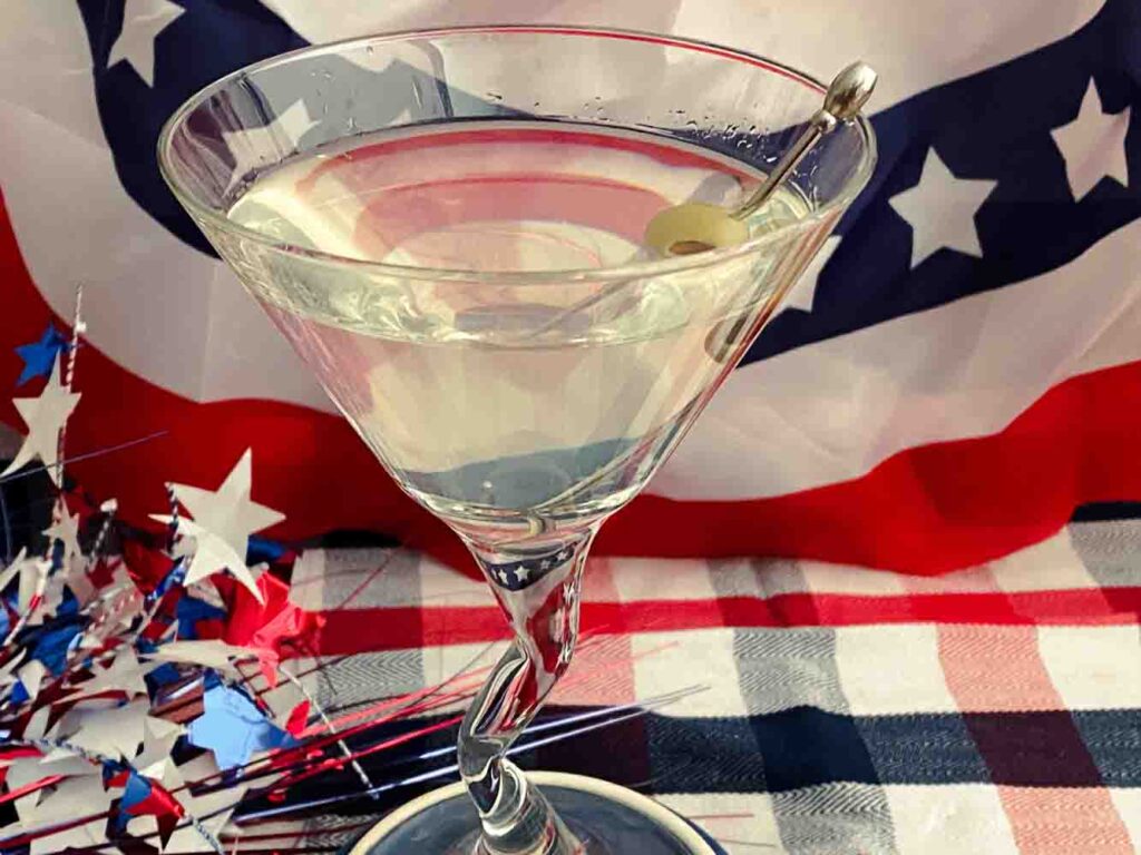 Top down picture of Absintini Ballot Cocktail with green olive in a Martini glass on top of red white blue checkered clothe. American flag background.