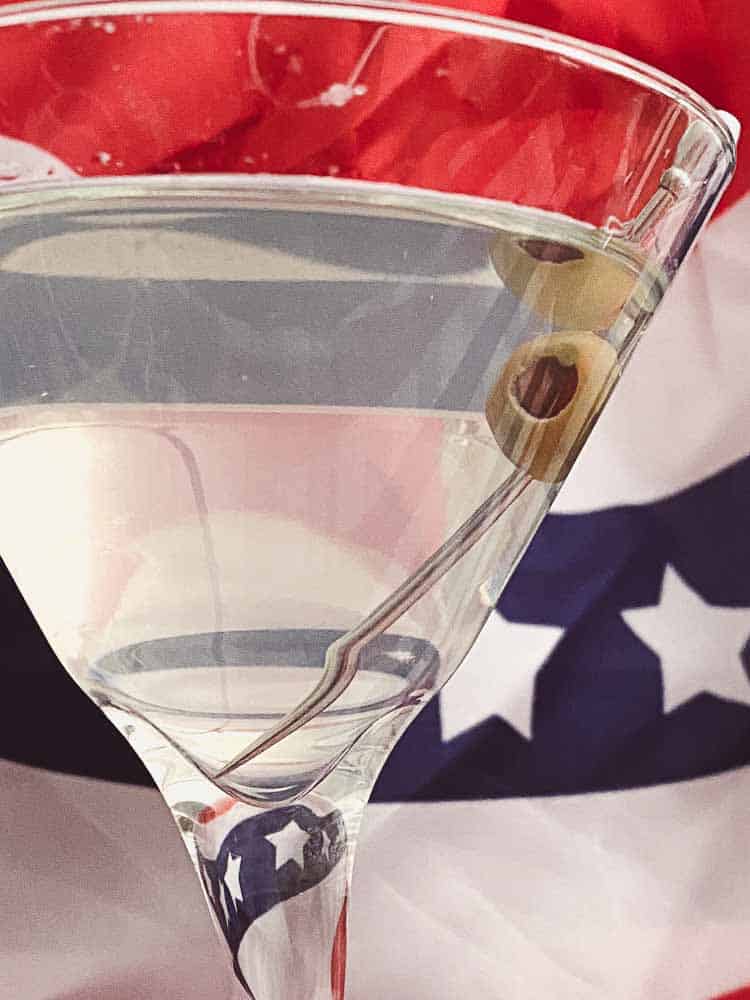 Absinthe Martini Cocktail close up with green olive and American flag decor in background.