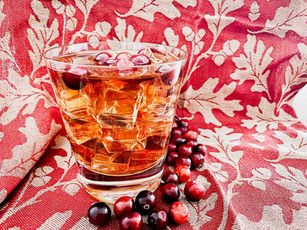 Cape Cod cocktail in a rocks glass with ice and garnished with fresh cranberries. Fresh cranberries wrapped around the bottle of the glass. Red and tan leaf clothe background.