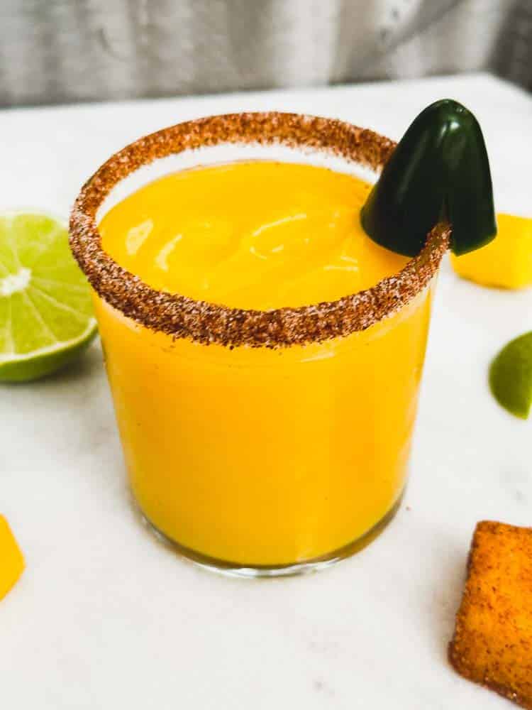 Chili Pepper Mango Margarita with Chili Pepper Salt rim and Sliced jalapeno garnish. slices of limes and mangoes in background.