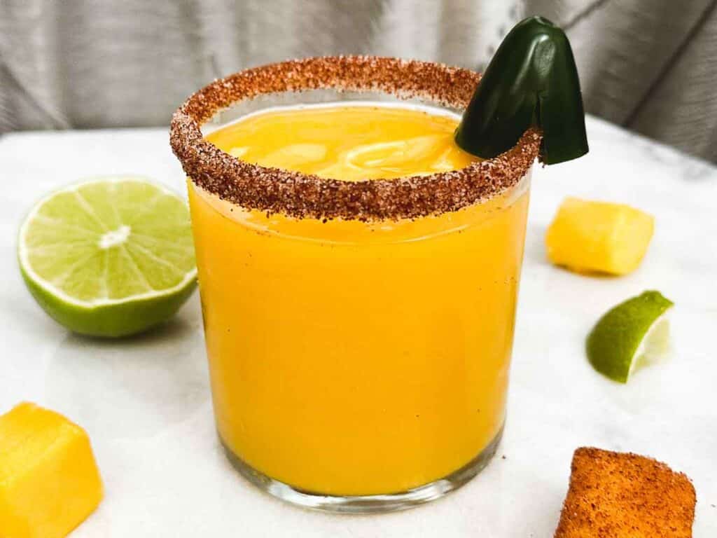 Chili Pepper Mango Margarita with Chili Pepper Salt rim and Sliced jalapeno garnish. slices of limes and mangoes in background.