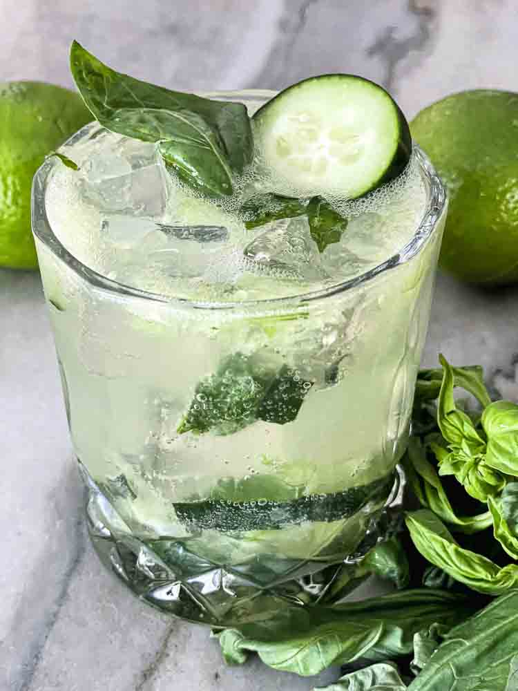 Cucumber Splash Mocktail in rocks glass. Cucumber garnish. Basil and Lime background.