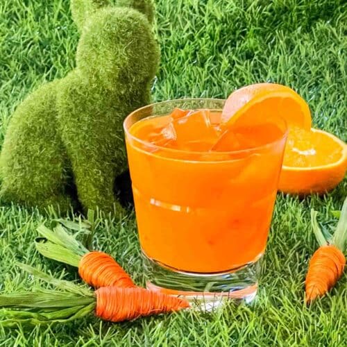 An orange Bunny Tonic Cocktail sitting on grass with little carrots and a green bunny around it