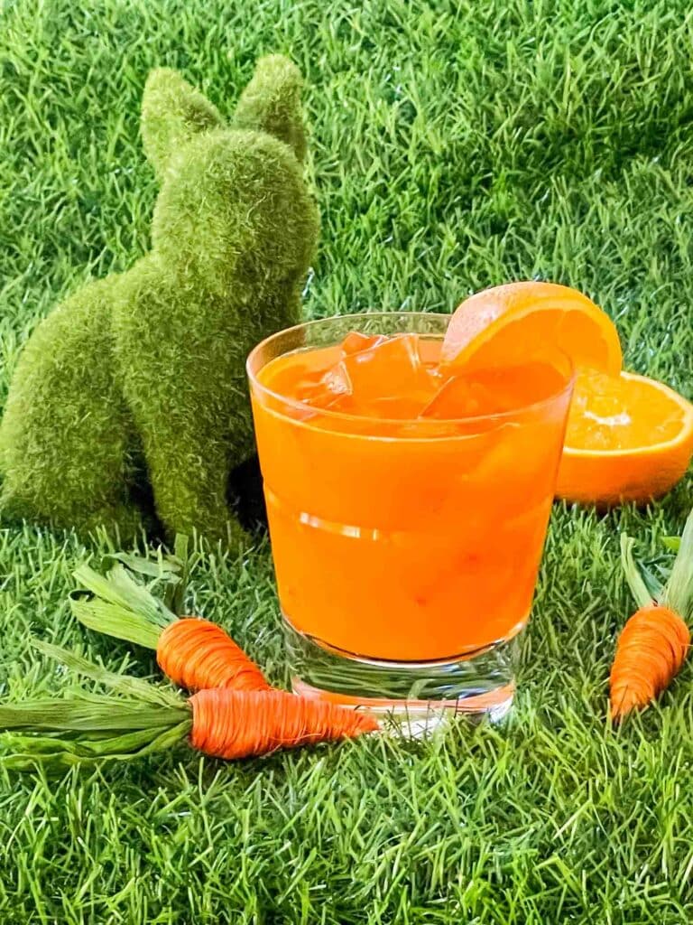 An orange Bunny Tonic Cocktail sitting on grass with little carrots and a green bunny around it