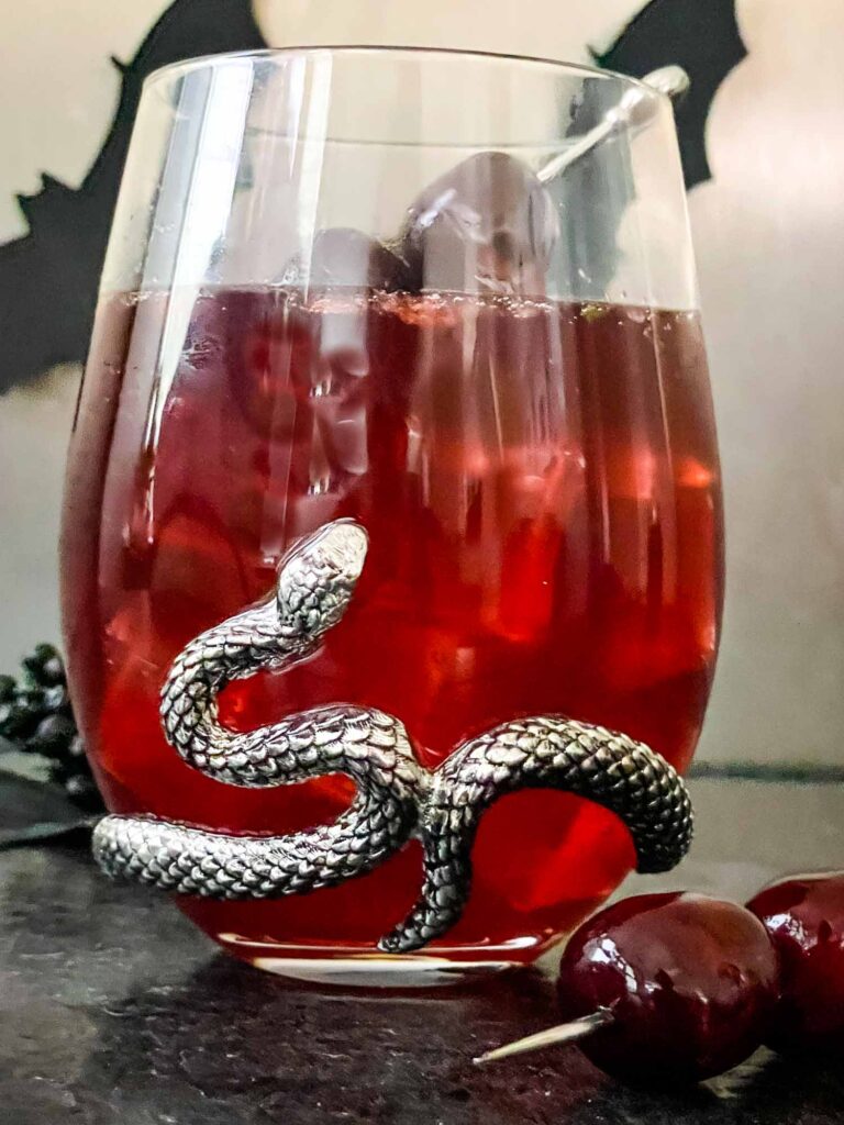 Close view of Bloody Heart Halloween Cocktail in a clear stemless glass with a silver snake. A red drink made with cherry and vodka.