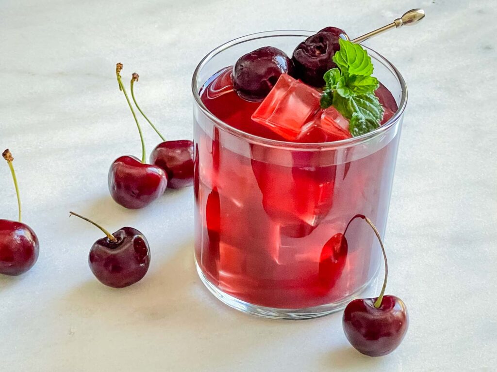 A glass of Cherry Vodka Cocktail with cherries around it on a white counter