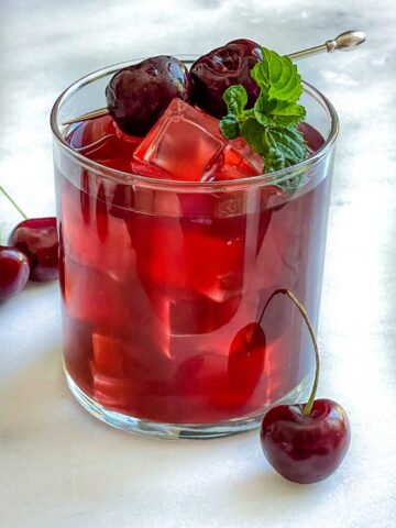 A glass of Cherry Vodka Cocktail with a mint sprig and brandied cherries on a white counter