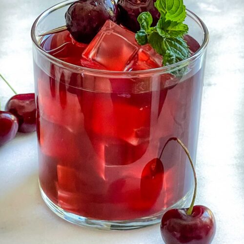 A glass of Cherry Vodka Cocktail with a mint sprig and brandied cherries on a white counter