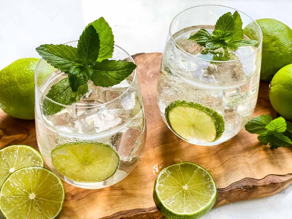 A wood board with two glasses of Hugo Spritz with fresh limes and mint sprigs