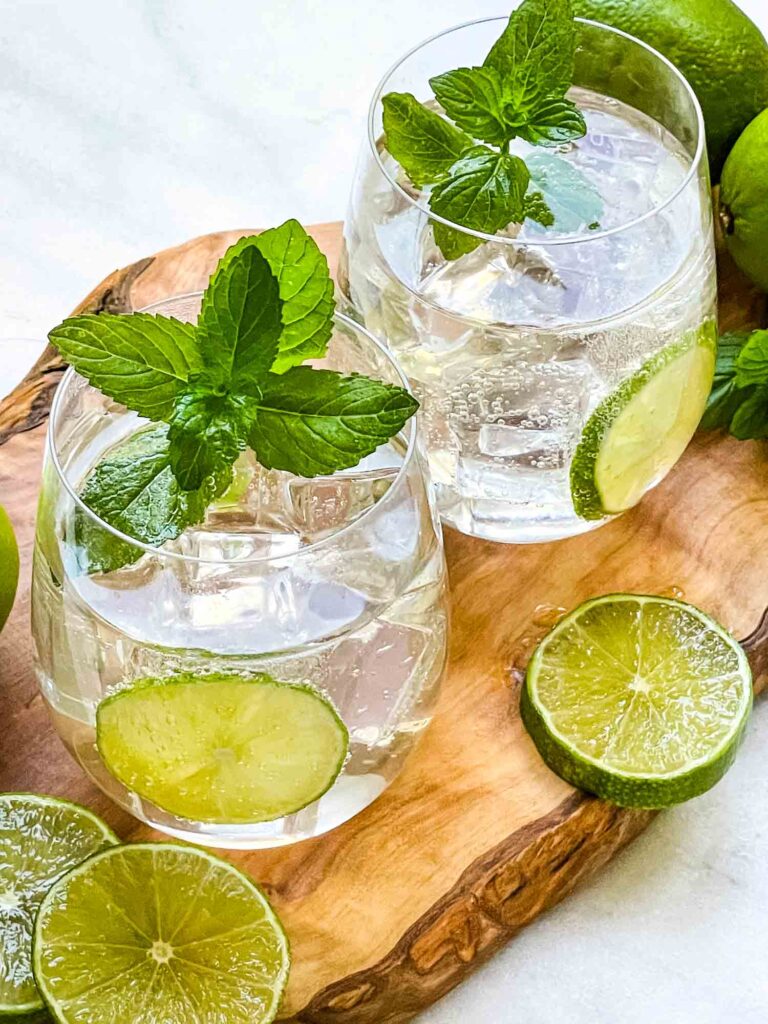Two clear glasses of Hugo Spritz garnished with mint and lime sitting on a wood board