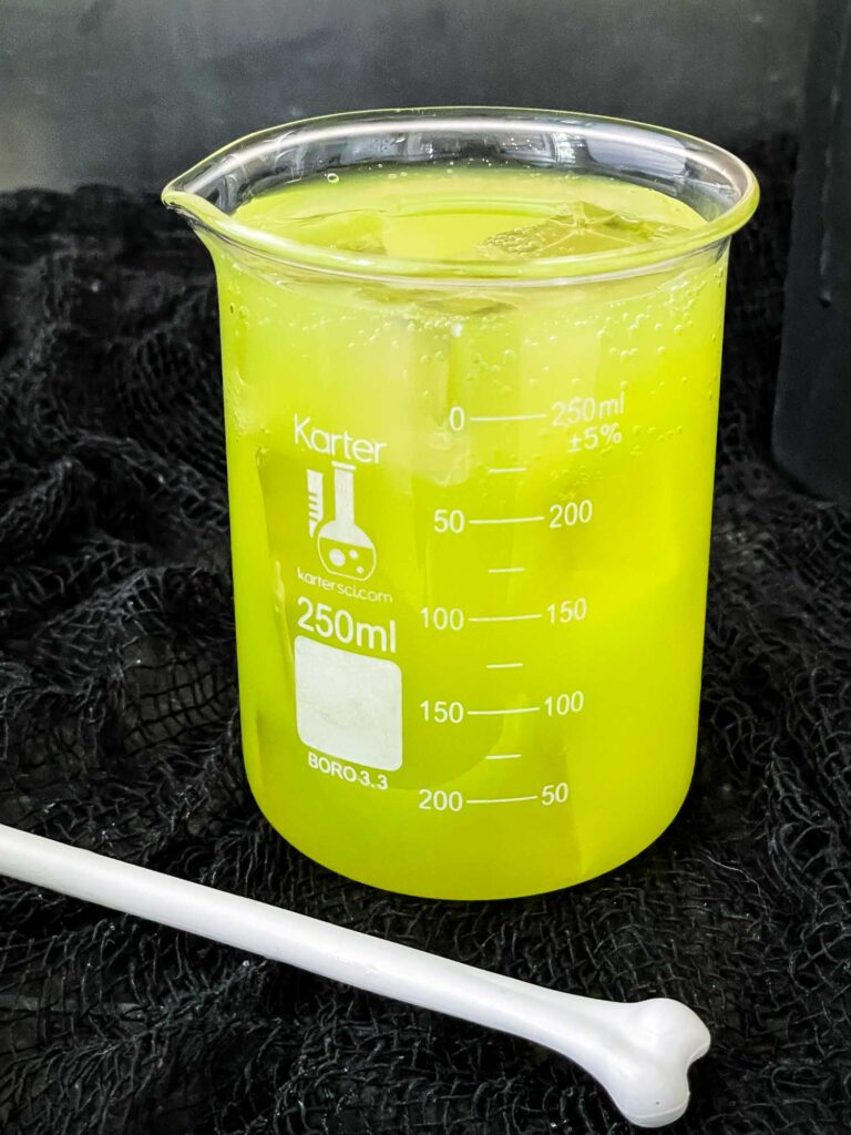 A bright green vodka Toxic Waste Cocktail in a beaker on a black cloth with a white bone stir stick