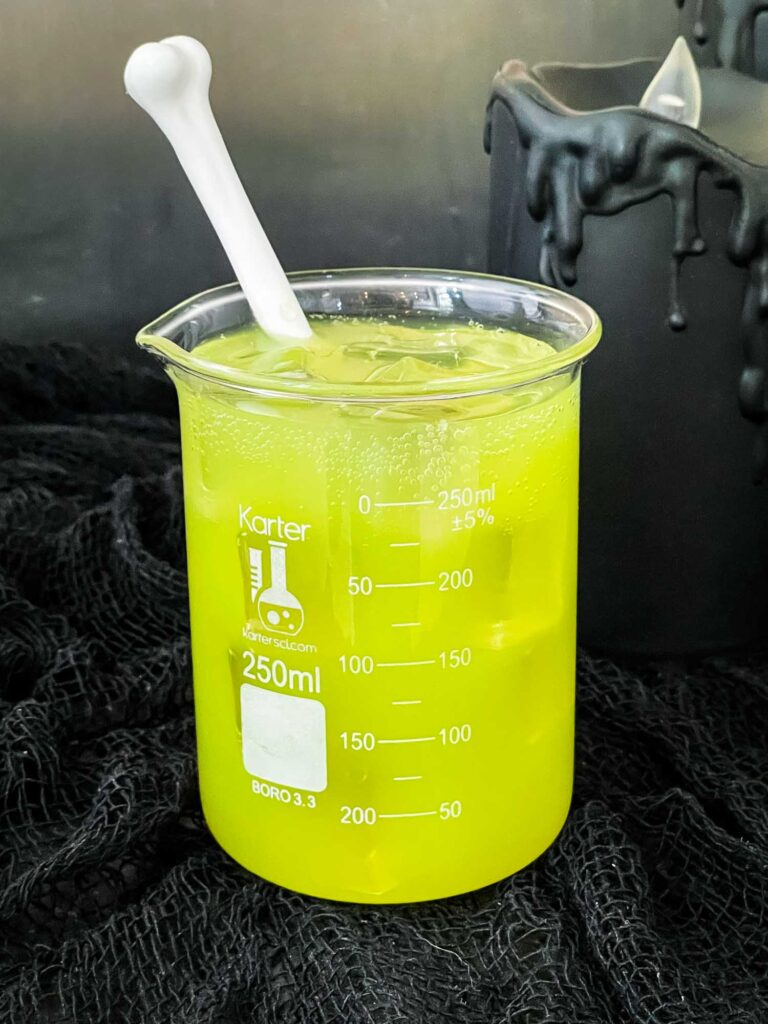 Bright green vodka Toxic Waste Cocktail in a glass beaker with a white bone stir stick on a black cloth