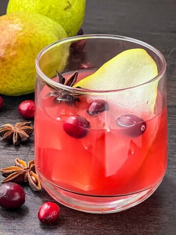 A glass full of Autumn Harvest Cocktail with floating cranberries and a pear slice sitting on a wood table