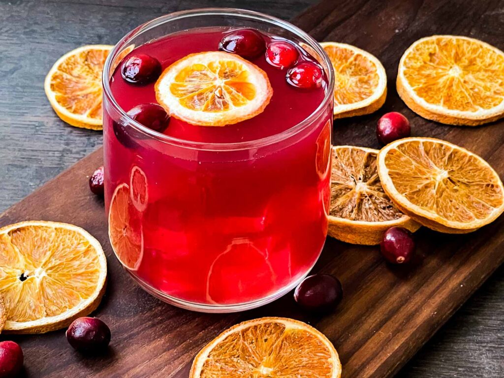 A glass of Cranberry Bog Cocktail on a wood board surrounded by dried orange slices and cranberries