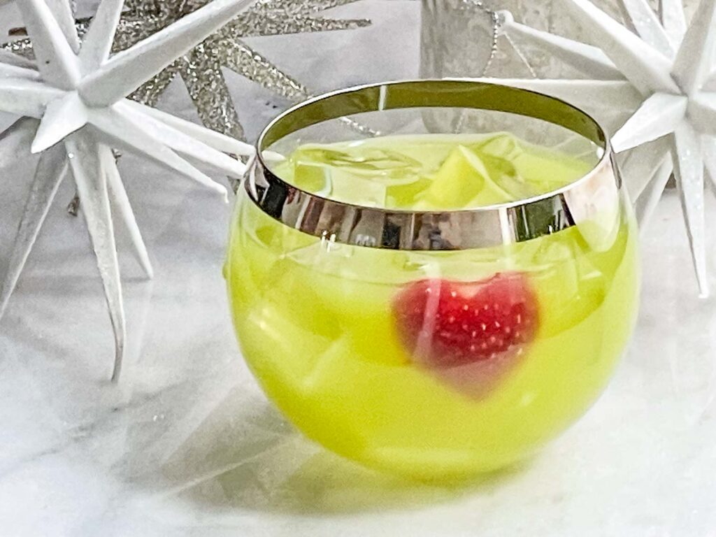 A round glass full of a green Grinch Cocktail with a red strawberry heart on a white counter