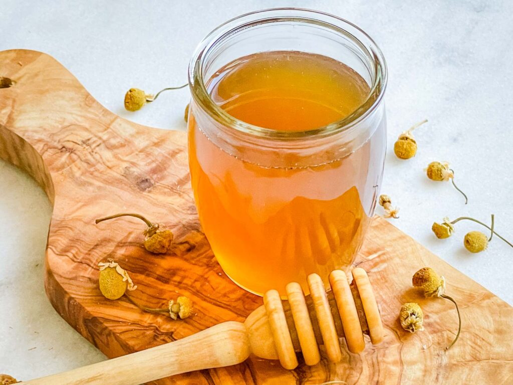 A glass jar of Honey Simple Syrup on a wood board with a honey stirrer