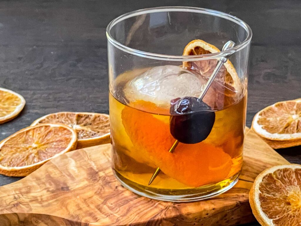 A glass of Bourbon Maple Old Fashioned on a wood board surrounded by dried orange slices.