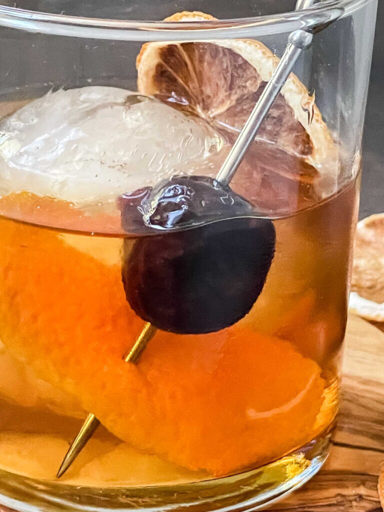 A close view of Bourbon Maple Old Fashioned in a glass with one large ice sphere, a brandied cherry, an orange peel, and dried orange slice.