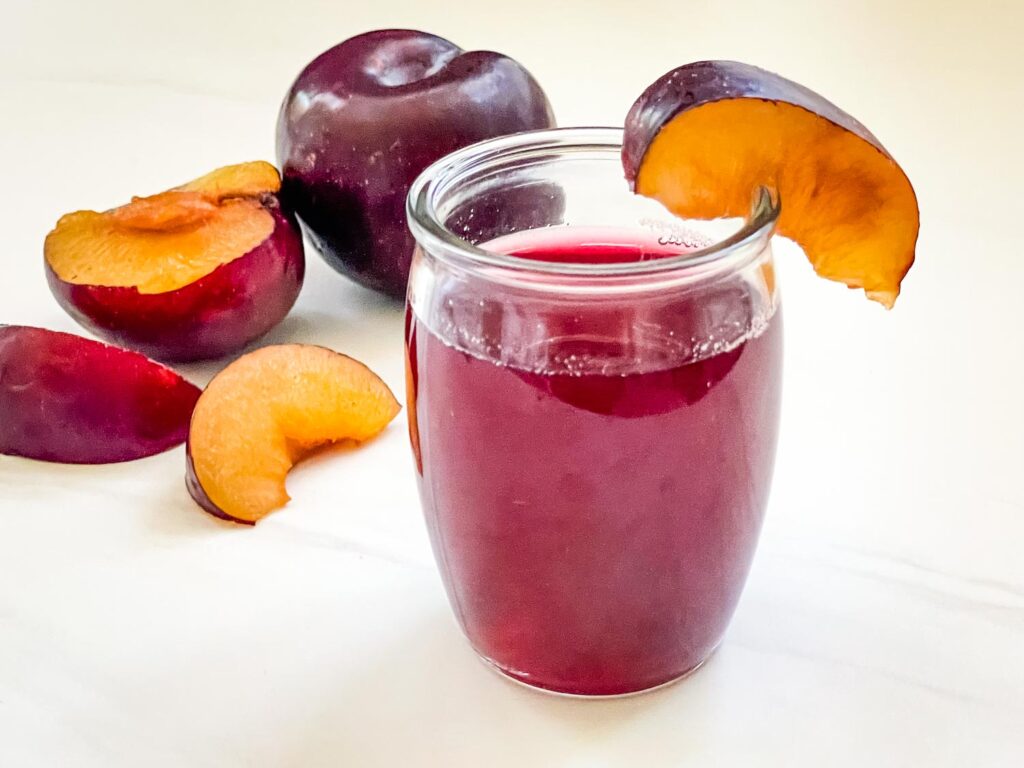 A small glass full of Plum Simple Syrup with fresh plums behind it on a white counter