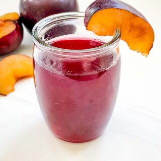 A small glass of Plum Simple Syrup on a white counter with fresh plums behind it