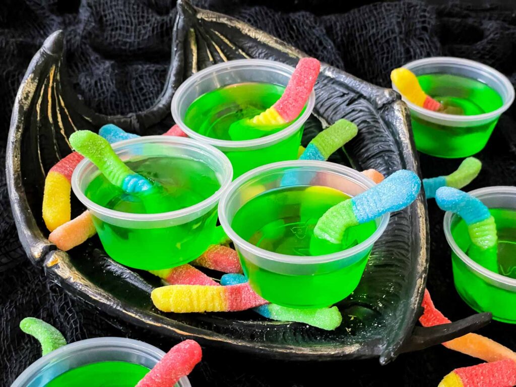 Cups of green Rotten Apple Halloween Jello Shots with gummy worms sitting on a bat dish