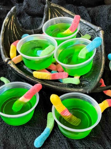 Cups of green Rotten Apple Halloween Jello Shots with gummy worms in them in a pile on a dish