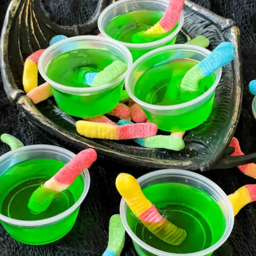 Cups of green Rotten Apple Halloween Jello Shots with gummy worms in them in a pile on a dish