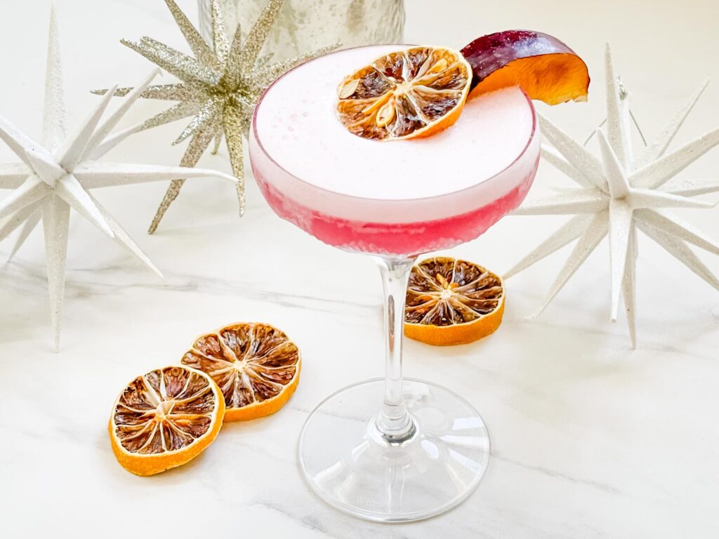 A Sugar Plum Fairy Cocktail (Plum Sour) in a glass coupe on a white counter with dried lemon wheels and decor behind it