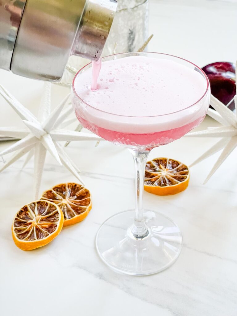 A silver cocktail shaker pouring Sugar Plum Fairy Cocktail (Plum Sour) into a glass coupe