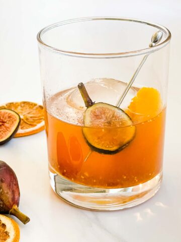 Fig Old Fashioned in a glass with ice on a white counter with a fresh fig inside.