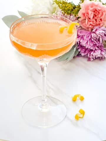 A glass of French Blonde Cocktail with a lemon twist sitting on a counter in front of flowers. Taylor Swift's favorite cocktail.