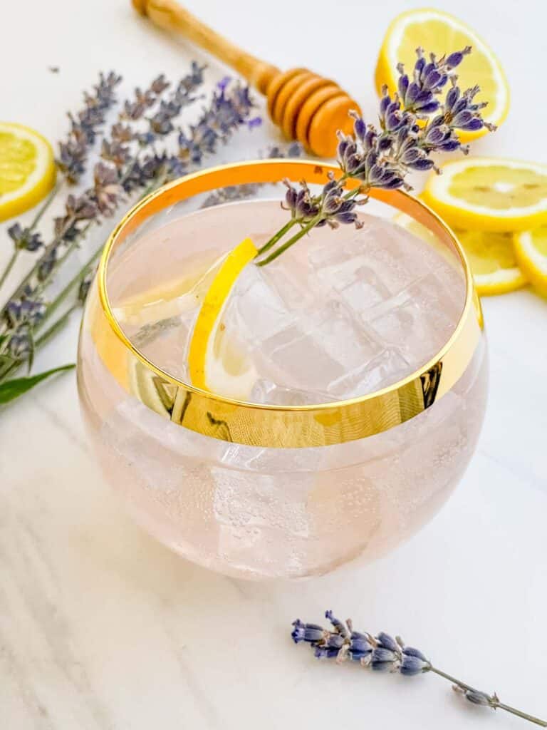 A glass of a Lavender Garden Cocktail garnished and surrounded by lavender and lemon pieces.