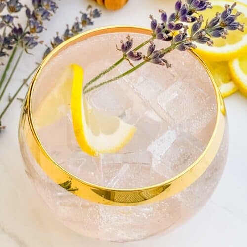 A glass with a gold rim full of a Lavender Garden Cocktail with a lemon wheel and lavender sprig in it.