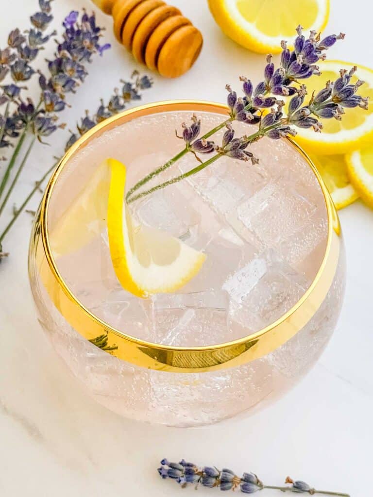 A glass with a gold rim full of a Lavender Garden Cocktail with a lemon wheel and lavender sprig in it.