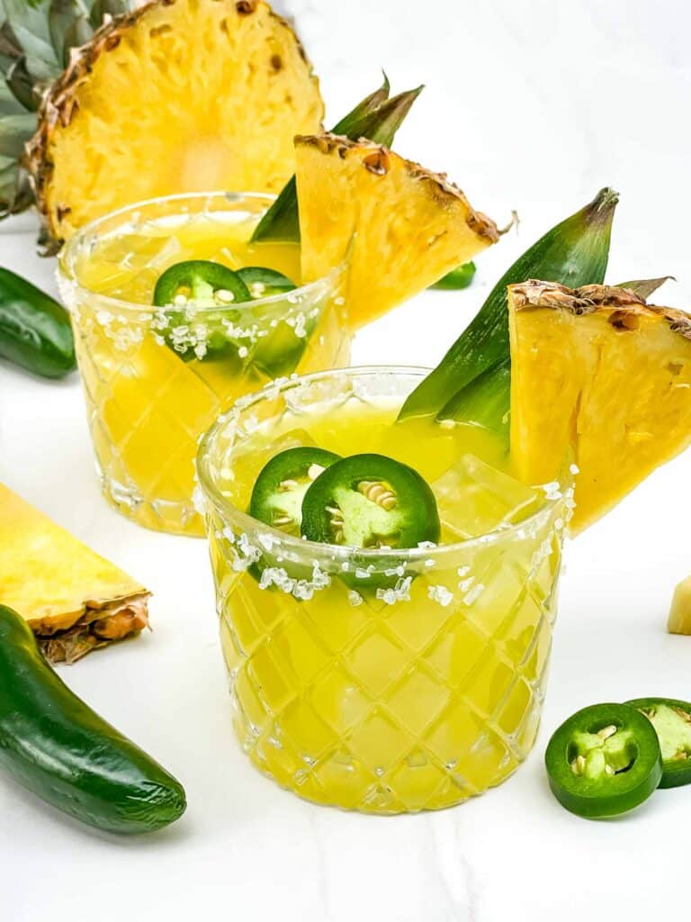 Two glasses of yellow Spicy Pineapple Mezcal Margarita on a counter with fresh garnishes.