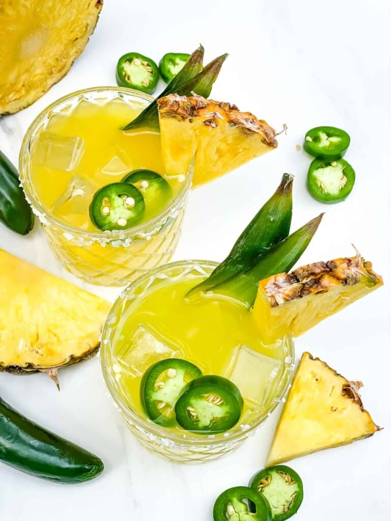 Looking down on two glasses of yellow Spicy Pineapple Mezcal Margarita on a counter with fresh ingredients around them.